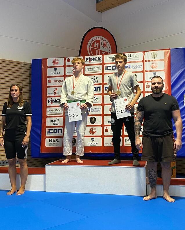 Drei Medaillenträger stehen auf dem Siegertreppchen nach einem Judowettbewerb, begleitet von einer Trainerin.