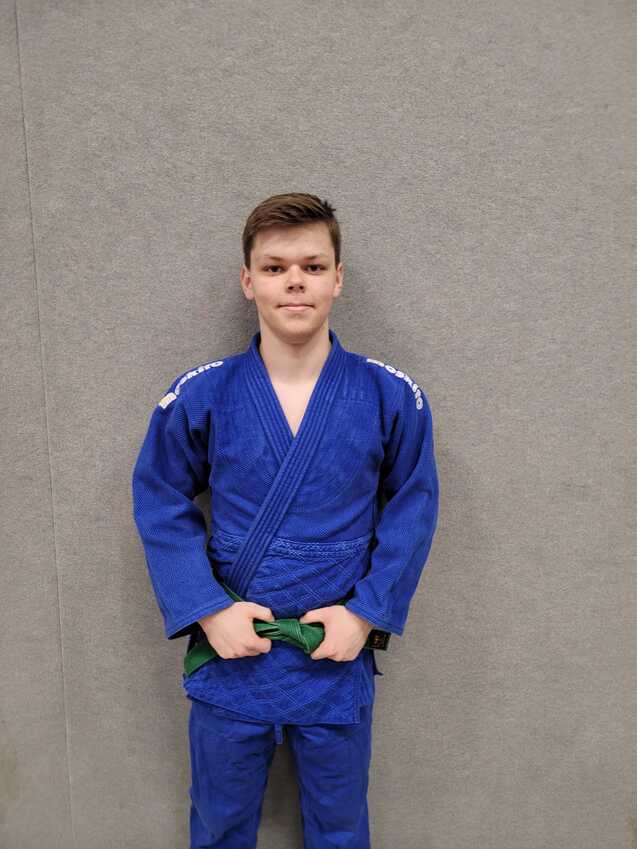 Junger Sportler in blauem Jiu-Jitsu-Anzug mit grünem Gürtel steht vor grauer Wand.