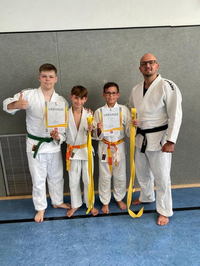 Vier Personen in Judo-Anzügen halten Urkunden und gelbe Gürtel, stehen vor einer grauen Wand und lächeln.