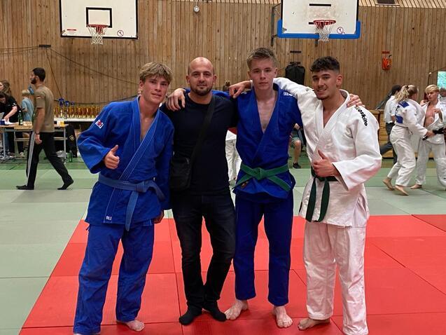 Vier Personen stehen lächelnd auf einer Judomatte, zwei in blauen und einer in weißem Gi, im Hintergrund andere Judoka.