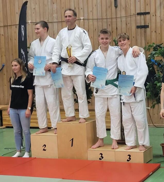 Vier Sportler in Judogi stehen auf einem Siegertreppchen, zwei halten Urkunden und der erste Platz hat einen Pokal.