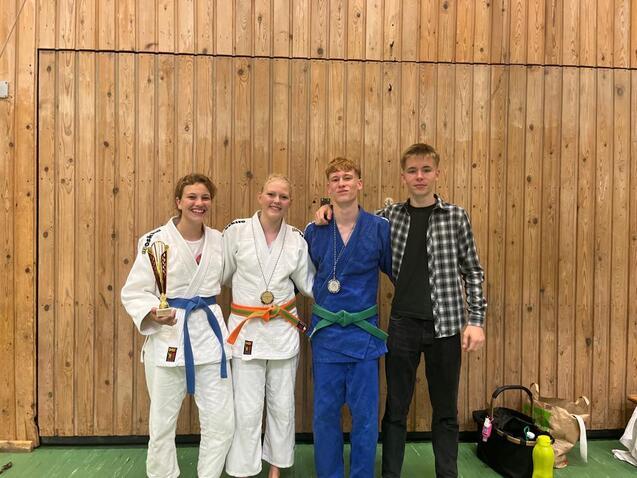 Vier Jugendliche posieren in einem Sportverein, zwei im Judoanzug mit Medaillen, eine mit Pokal, eine in Zivilkleidung.