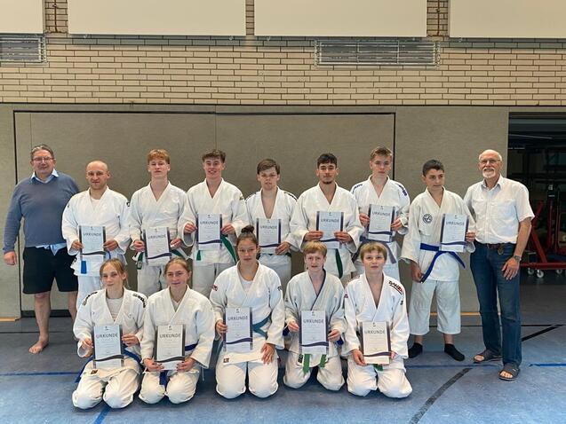 Gruppenfoto von Judoka in weißen Anzügen mit Urkunden, Stehend und Sitzend in einer Sporthalle.