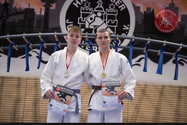 Zwei Judokas in weißen Anzügen posieren mit Medaillen und Urkunden nach einem Wettkampf in einer Sporthalle.