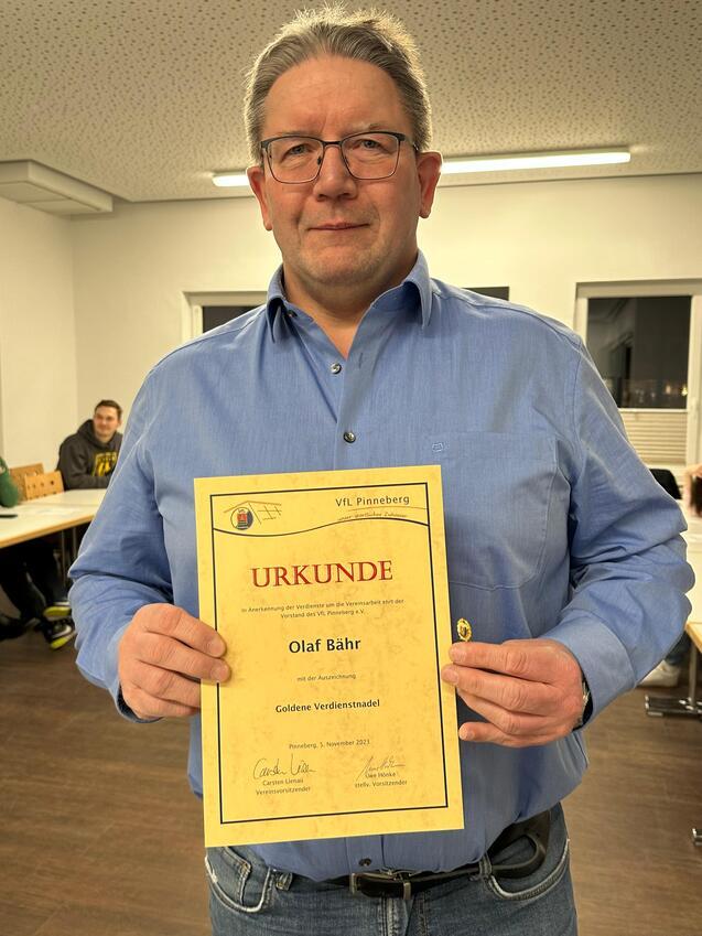 Mann in blauer Hemd hält eine Urkunde mit der Aufschrift „Goldene Verdienstnadel“ in einem Vereinsraum des VfL Pinneberg.