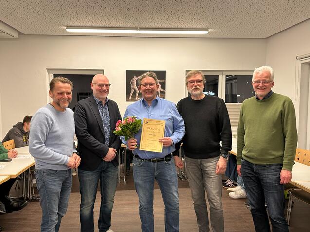 Gruppenfoto von fünf Männern, einer hält eine Urkunde und einen Blumenstrauß, in einem Raum mit Tischen.