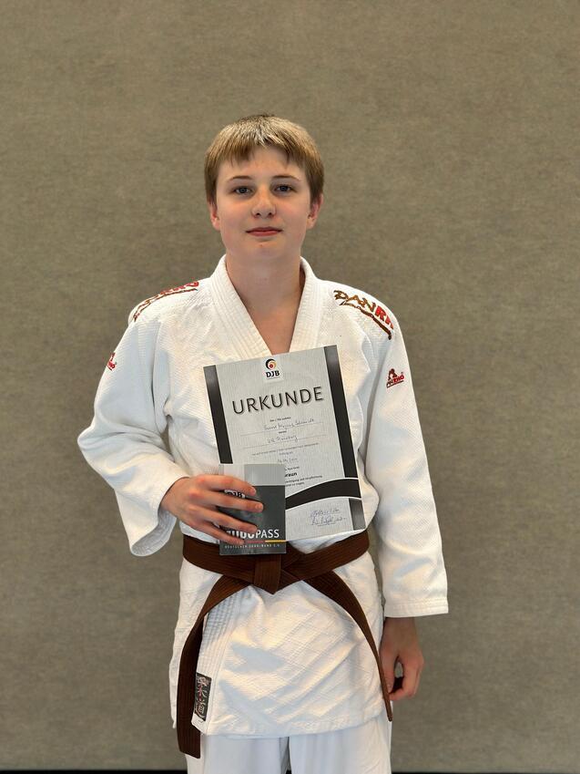 Junge im Judoanzug hält stolz eine Urkunde in der Hand, im Hintergrund eine neutrale Wand.