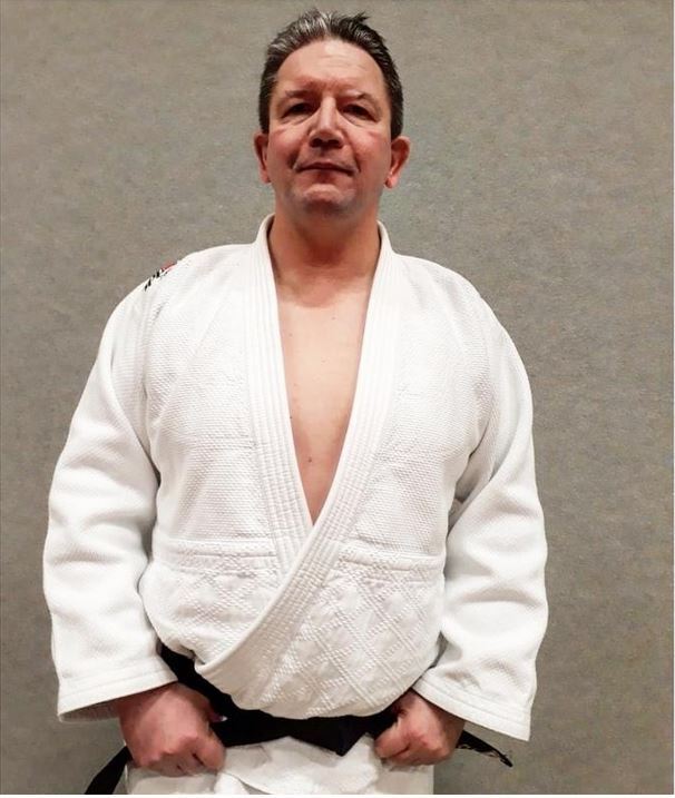 Mann in einem weißen Judoanzug mit schwarzem Gürtel posiert vor grauem Hintergrund.