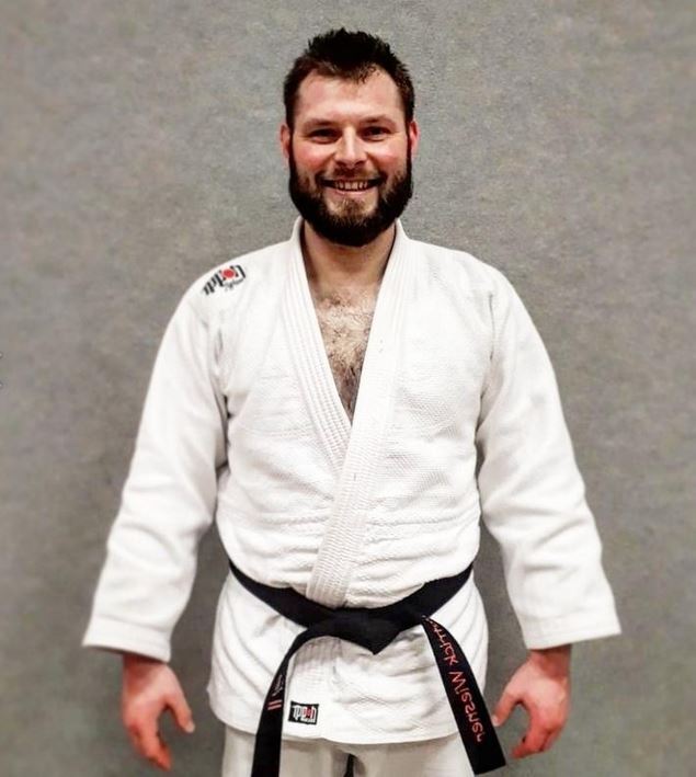 Ein lächelnder Mann in einem weißen Judoanzug mit schwarzem Gürtel steht vor grauem Hintergrund.