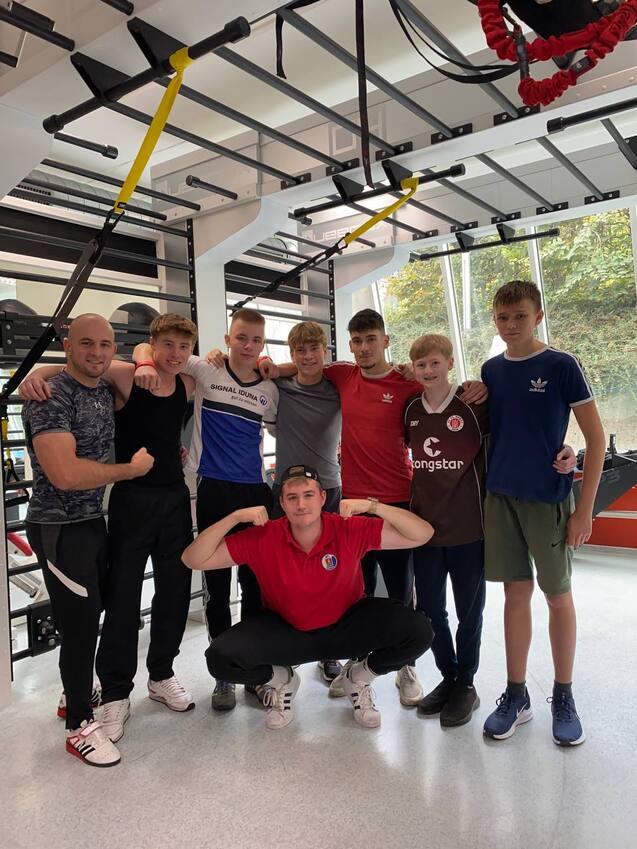 Gruppe von sieben Jugendlichen und einem Trainer in einem modernen Fitnessraum, lächelnd und posierend für das Foto.