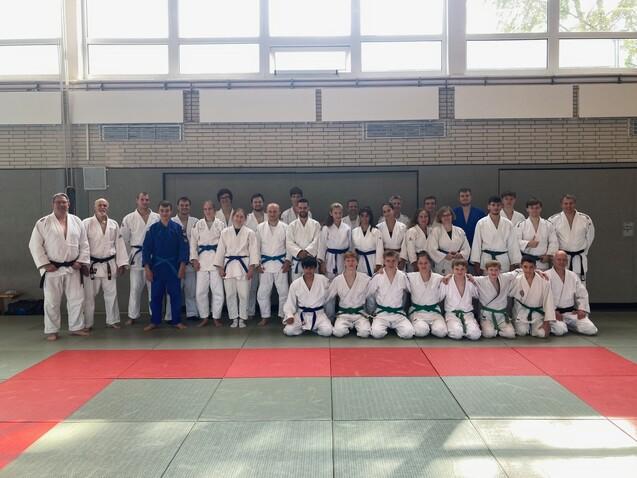 Gruppenfoto von Judoka in weißen und blauen Anzügen auf einer Matte in einem Trainingsraum.
