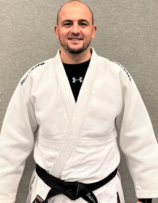 Mann im weißen Judoanzug mit schwarzem Gürtel, lächelt vor grauem Hintergrund.