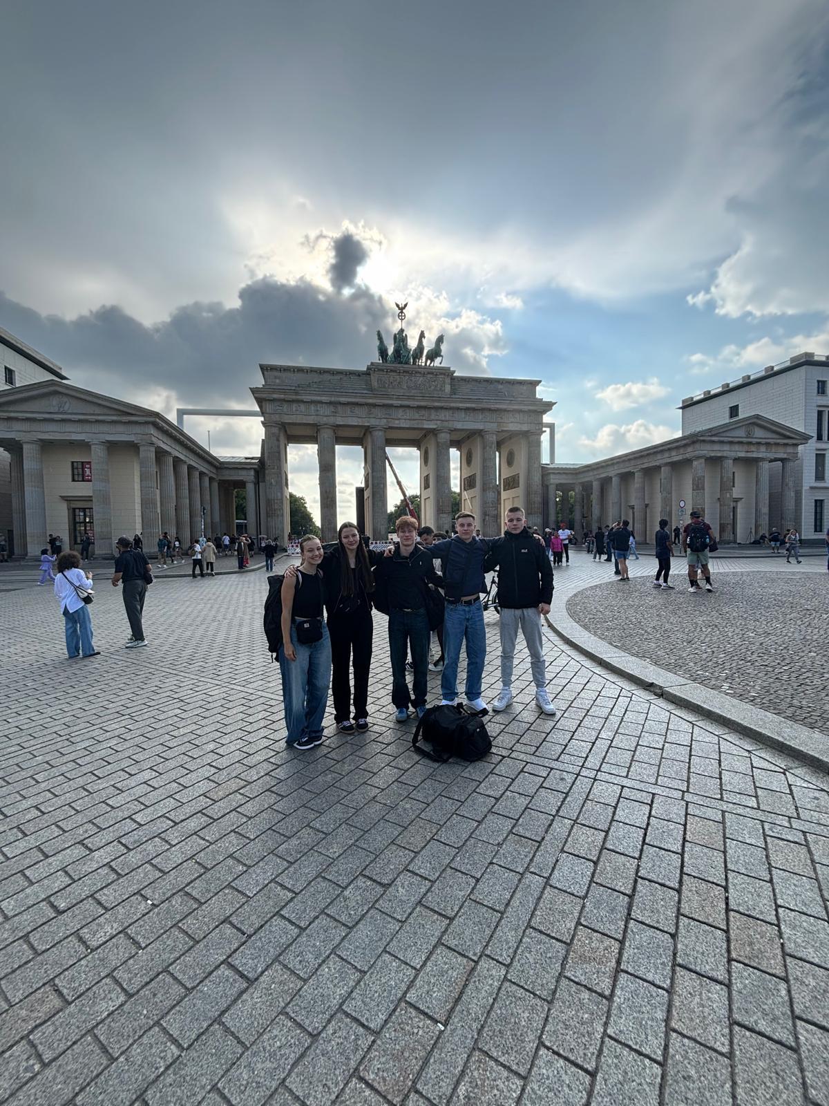 Gruppe von fünf Personen posiert vor dem Brandenburger Tor, umgeben von Besuchern auf einem gepflasterten Platz.