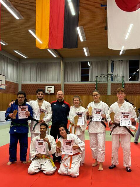 Gruppe von Judokas in weißen Anzügen mit Urkunden in einer Sporthalle, im Hintergrund hängen Flaggen verschiedener Länder.