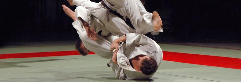 Zwei Judoka in weißen Anzügen führen einen Wurf auf einer Tatami mit rotem Rand aus.