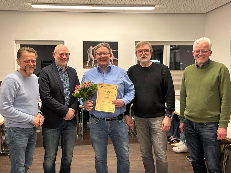 Gruppenfoto von fünf Männern, einer hält eine Urkunde und einen Blumenstrauß, in einem Raum mit Tischen.