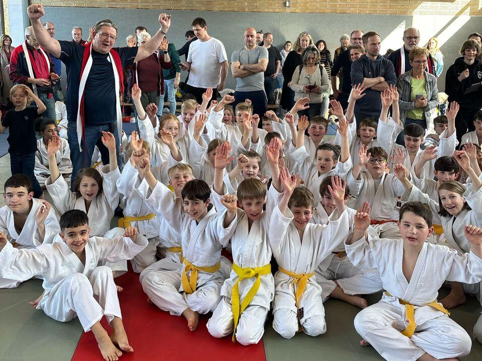 Gruppenselfie von Kindern in weißen Judojacken mit gelben Gürteln, umgeben von Zuschauern in einer Sporthalle.