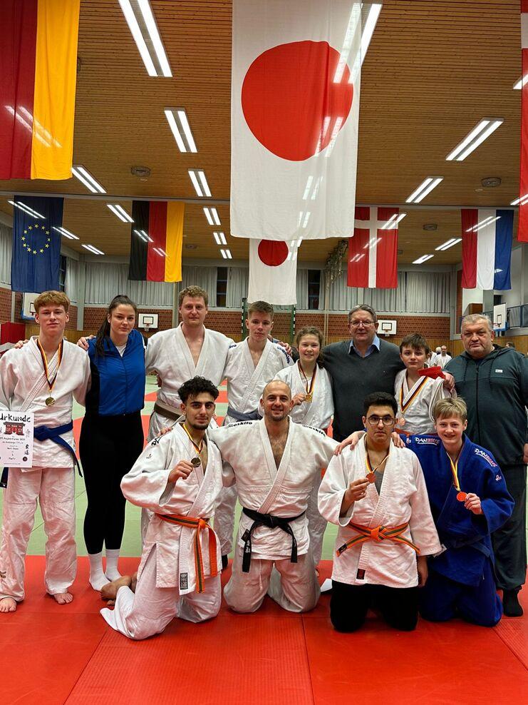 Gruppe von Judoka mit Medaillen in der Sporthalle, umgeben von Flaggen und Matten. Urkunden in Händen eines Teilnehmers.