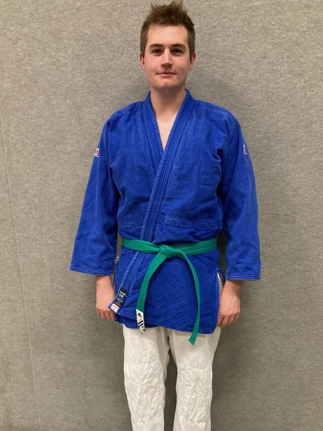 Junger Mann in blauer Judo-Jacke und grüner Gürtelschnalle, steht auf einer Matte vor einer grauen Wand.