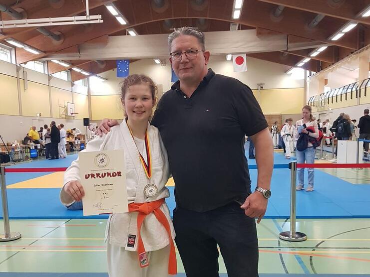 Junge in Karateanzug mit Medaille und Urkunde steht neben einem Mann in Sportbekleidung in einer Sporthalle.