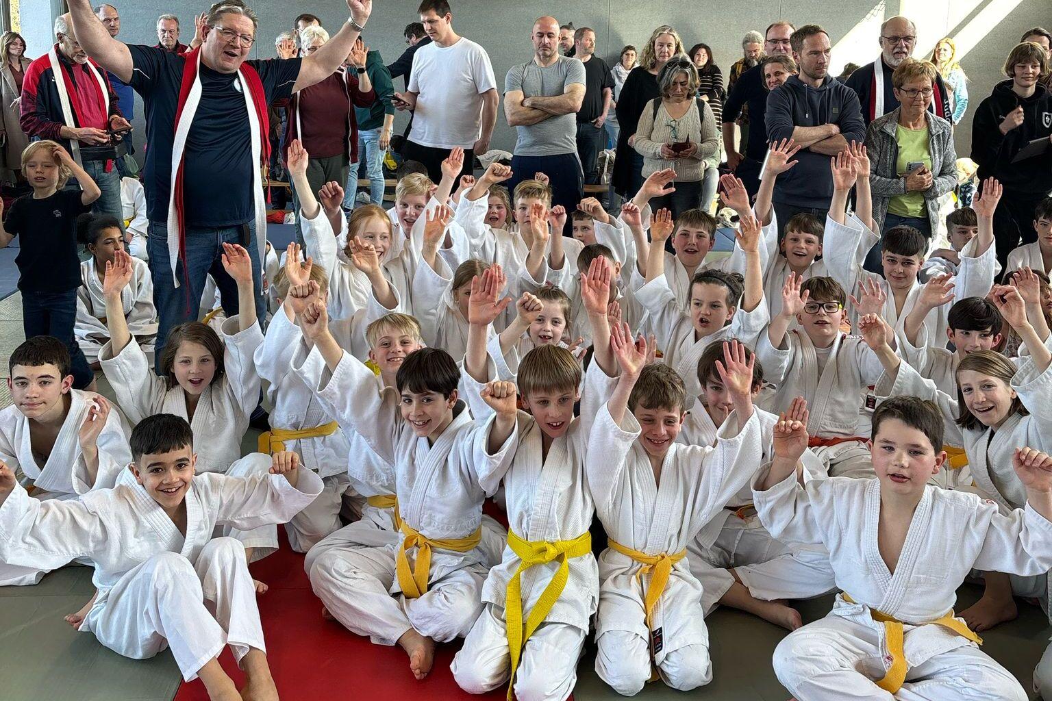 Gruppenselfie von Kindern in wei&szlig;en Judojacken mit gelben G&uuml;rteln, umgeben von Zuschauern in einer Sporthalle.