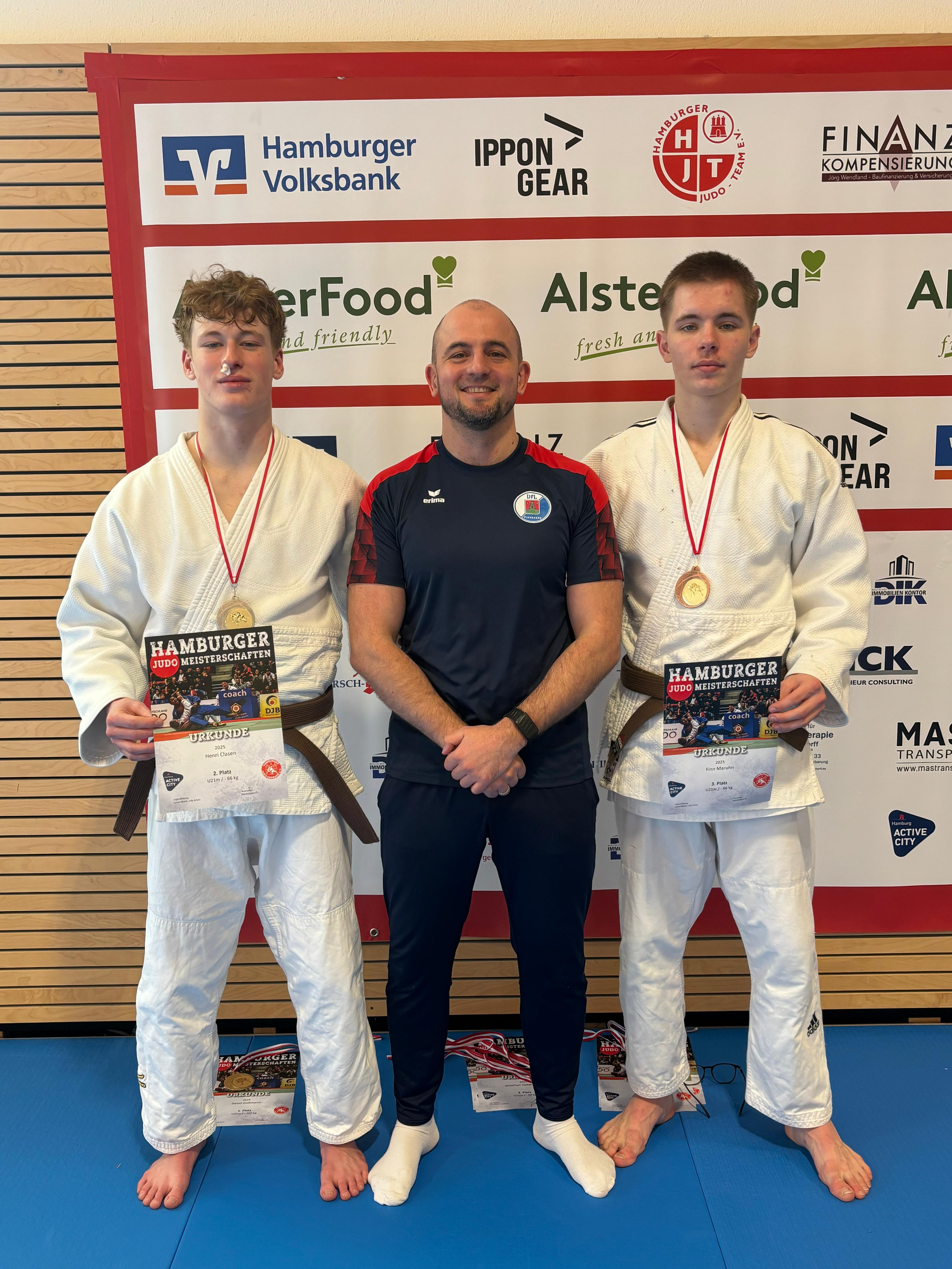 Zwei Judoka in weißen Anzügen und Medaillen stehen neben ihrem Trainer, der ein Zertifikat hält, vor einer Sponsorenwand.