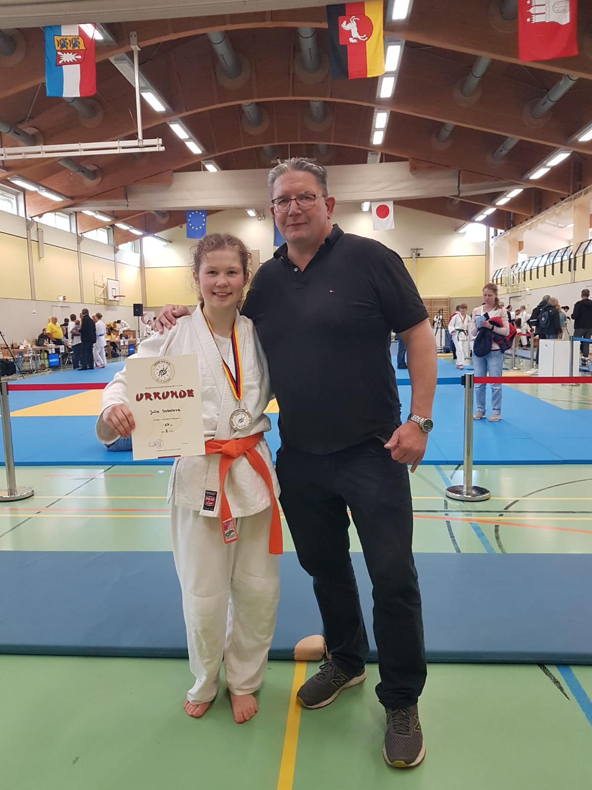 Junge in Karateanzug mit Medaille und Urkunde steht neben einem Mann in Sportbekleidung in einer Sporthalle.