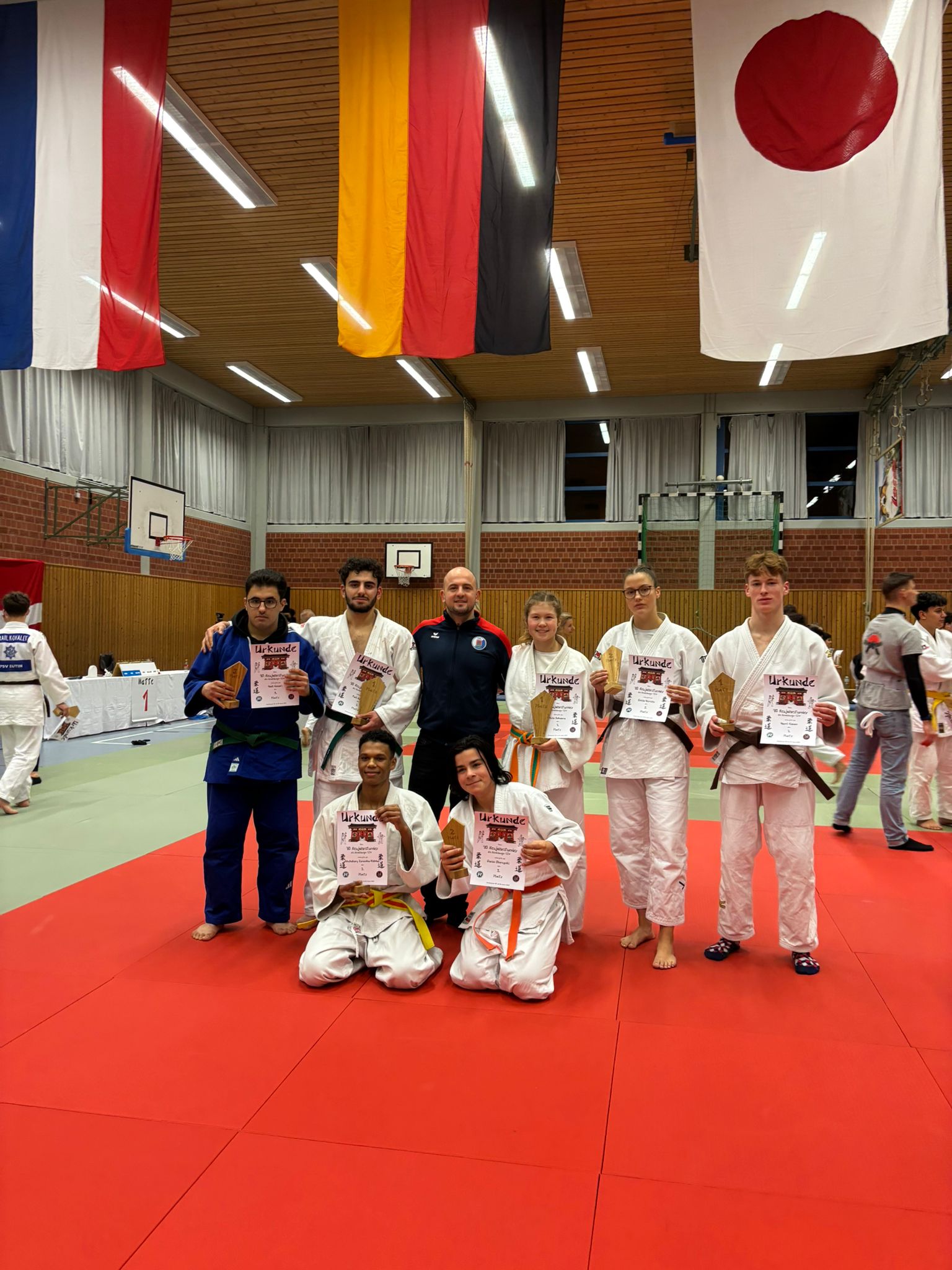 Gruppe von Judokas in weißen Anzügen mit Urkunden in einer Sporthalle, im Hintergrund hängen Flaggen verschiedener Länder.
