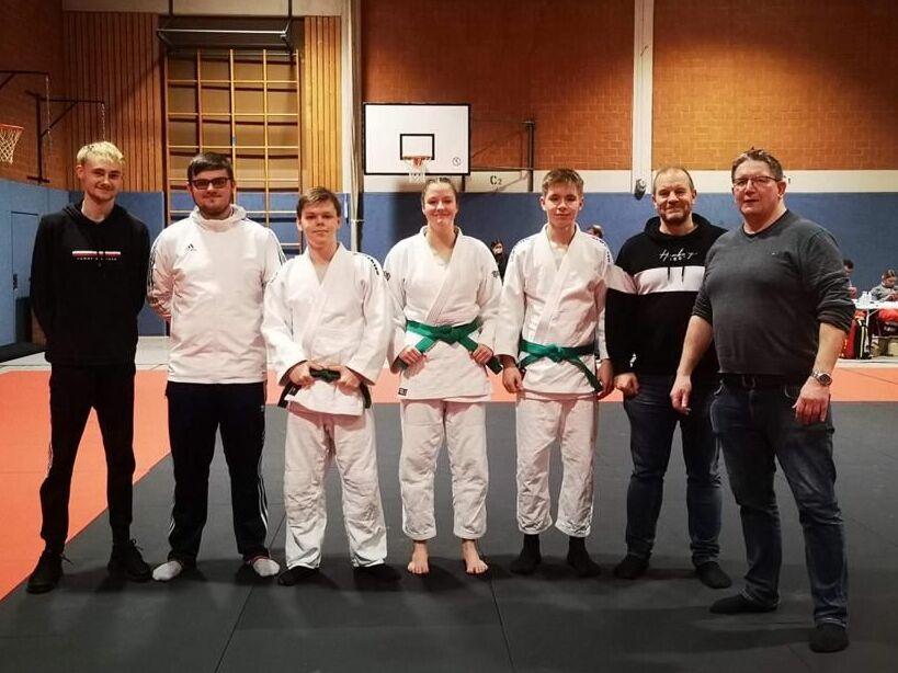 Gruppe von sechs Personen auf einer Judomatte; drei tragen Judoanzüge, zwei haben grüne Gürtel.
