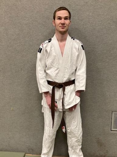 Junger Mann in einem wei&szlig;en Judogi mit braunem G&uuml;rtel steht auf einer Tatami. Hintergrund ist eine graue Wand.