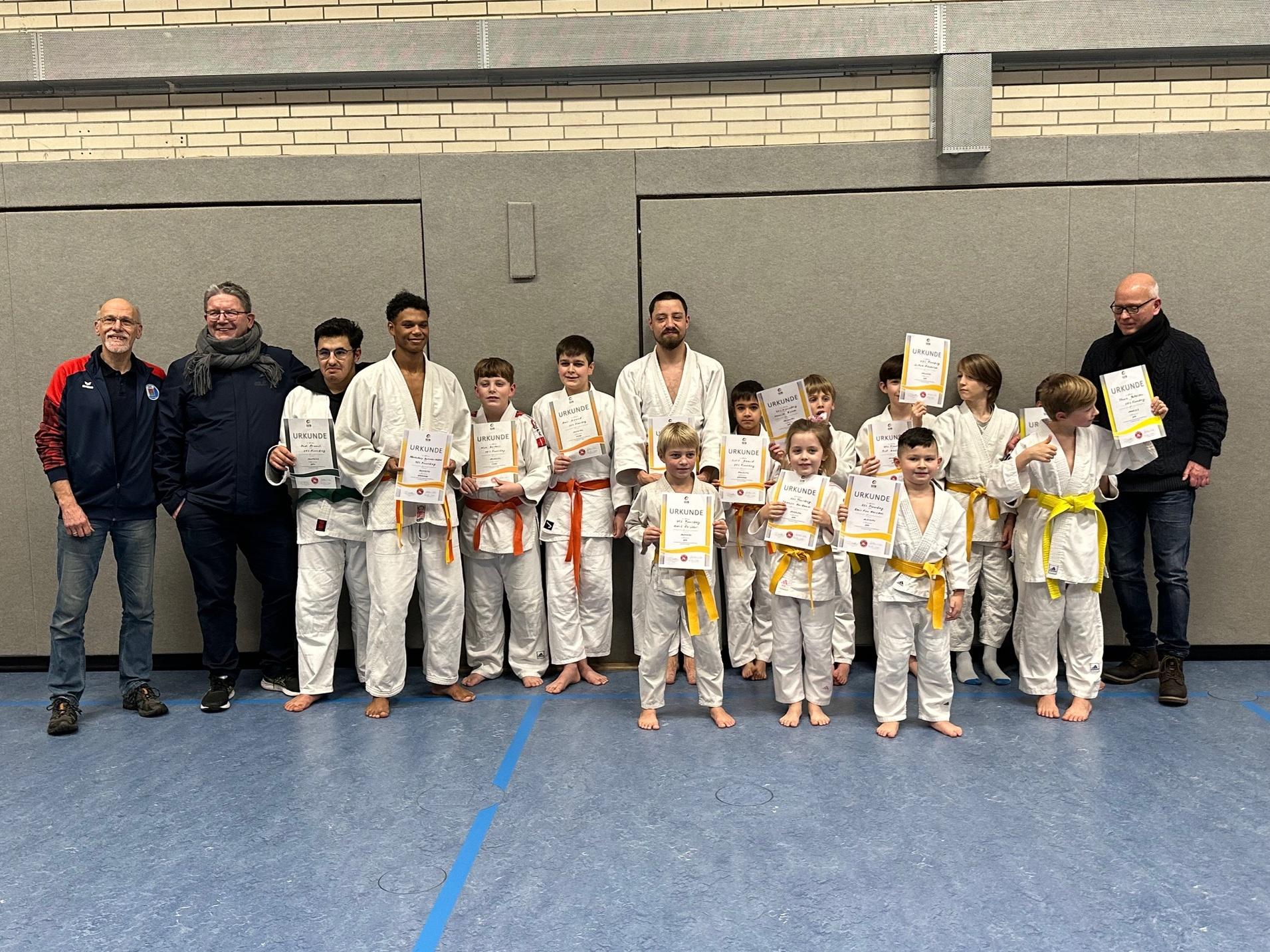 Gruppennfoto von Kindern und Trainern in weißen Judoanzügen, jeder mit Urkunden in der Hand, in einer Sporthalle.