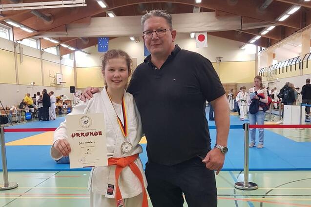 Junge in Karateanzug mit Medaille und Urkunde steht neben einem Mann in Sportbekleidung in einer Sporthalle.