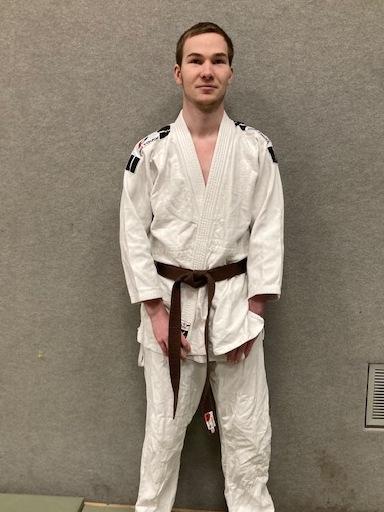 Junger Mann in einem weißen Judogi mit braunem Gürtel steht auf einer Tatami. Hintergrund ist eine graue Wand.