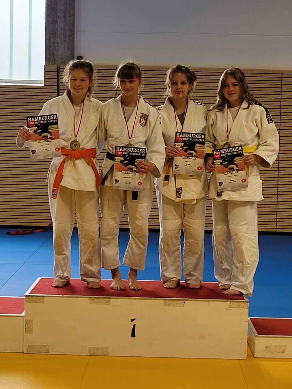 Vier Judoka auf dem Siegertreppchen halten Medaillen und Urkunden nach einem Wettkampf in einer Turnhalle.