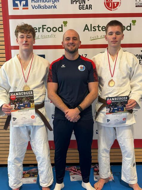 Zwei Judoka in weißen Anzügen und Medaillen stehen neben ihrem Trainer, der ein Zertifikat hält, vor einer Sponsorenwand.