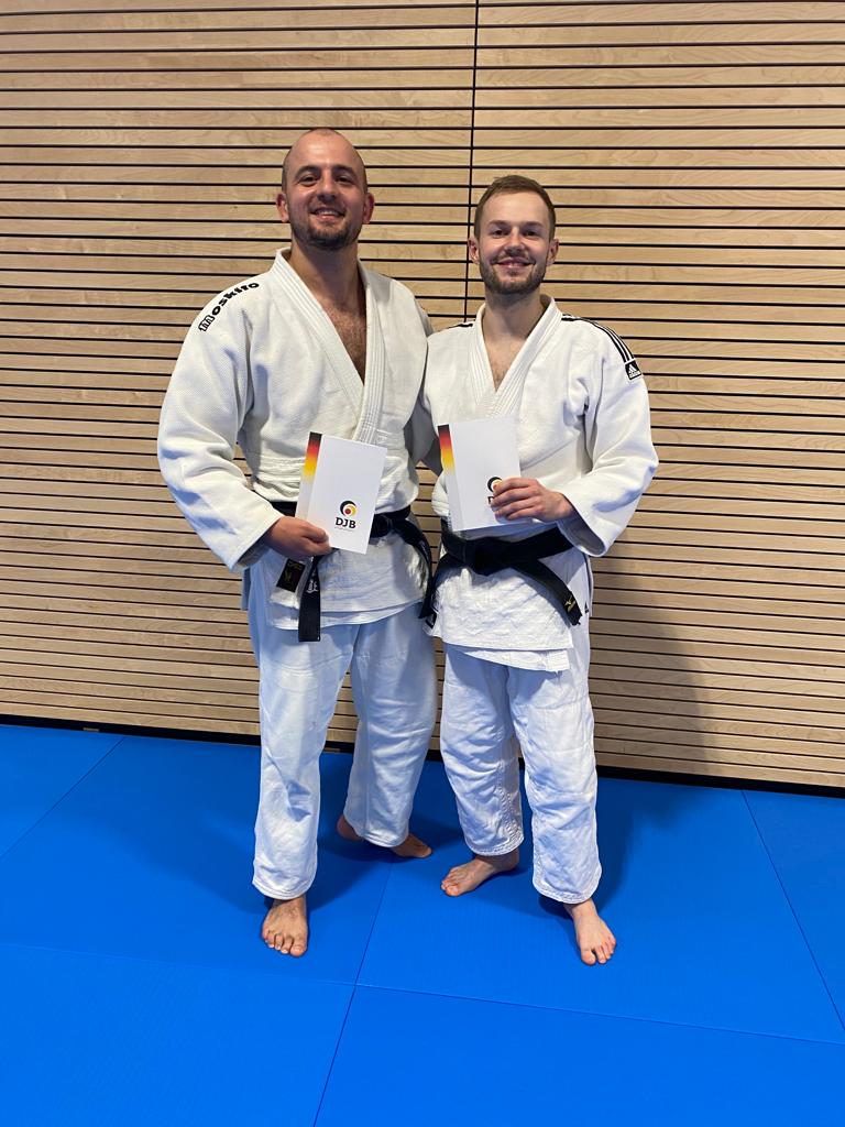 Zwei Judoka in weißem Gi mit schwarzen Gürtel halten Urkunden in der Hand und stehen auf einer blauen Matte vor Holzblenden.