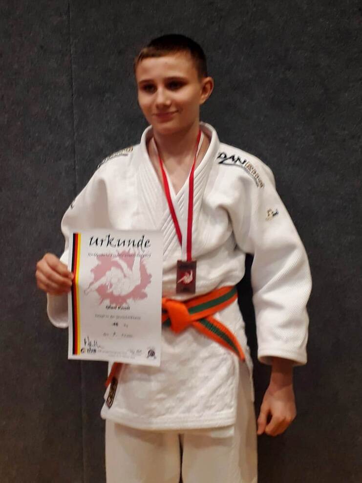 Ein junger Judoka in weißem Gi hält stolz eine Urkunde und eine Medaille vor einem grauen Hintergrund.