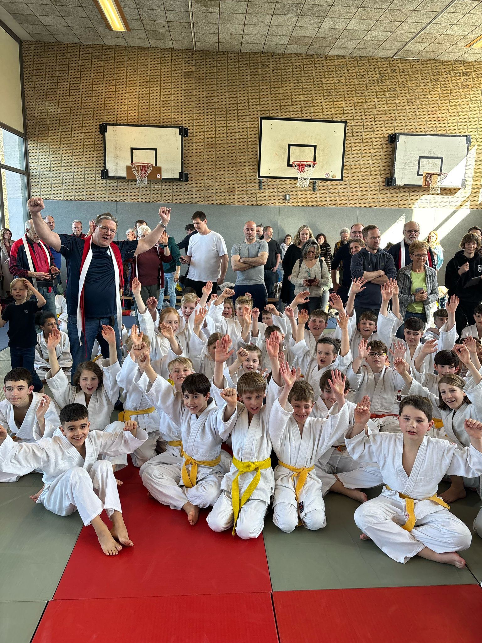 Gruppenselfie von Kindern in weißen Judojacken mit gelben Gürteln, umgeben von Zuschauern in einer Sporthalle.