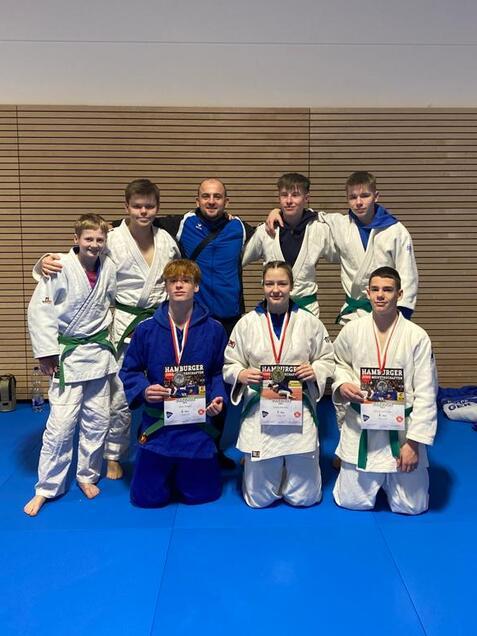 Gruppe junger Judoka in Traininganz&uuml;gen mit Medaillen und Urkunden auf einer blauen Matte in einem Sportraum.