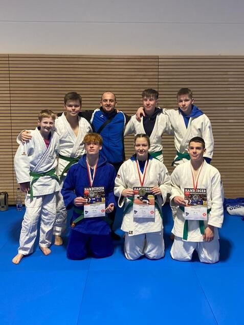 Gruppe junger Judoka in Traininganzügen mit Medaillen und Urkunden auf einer blauen Matte in einem Sportraum.