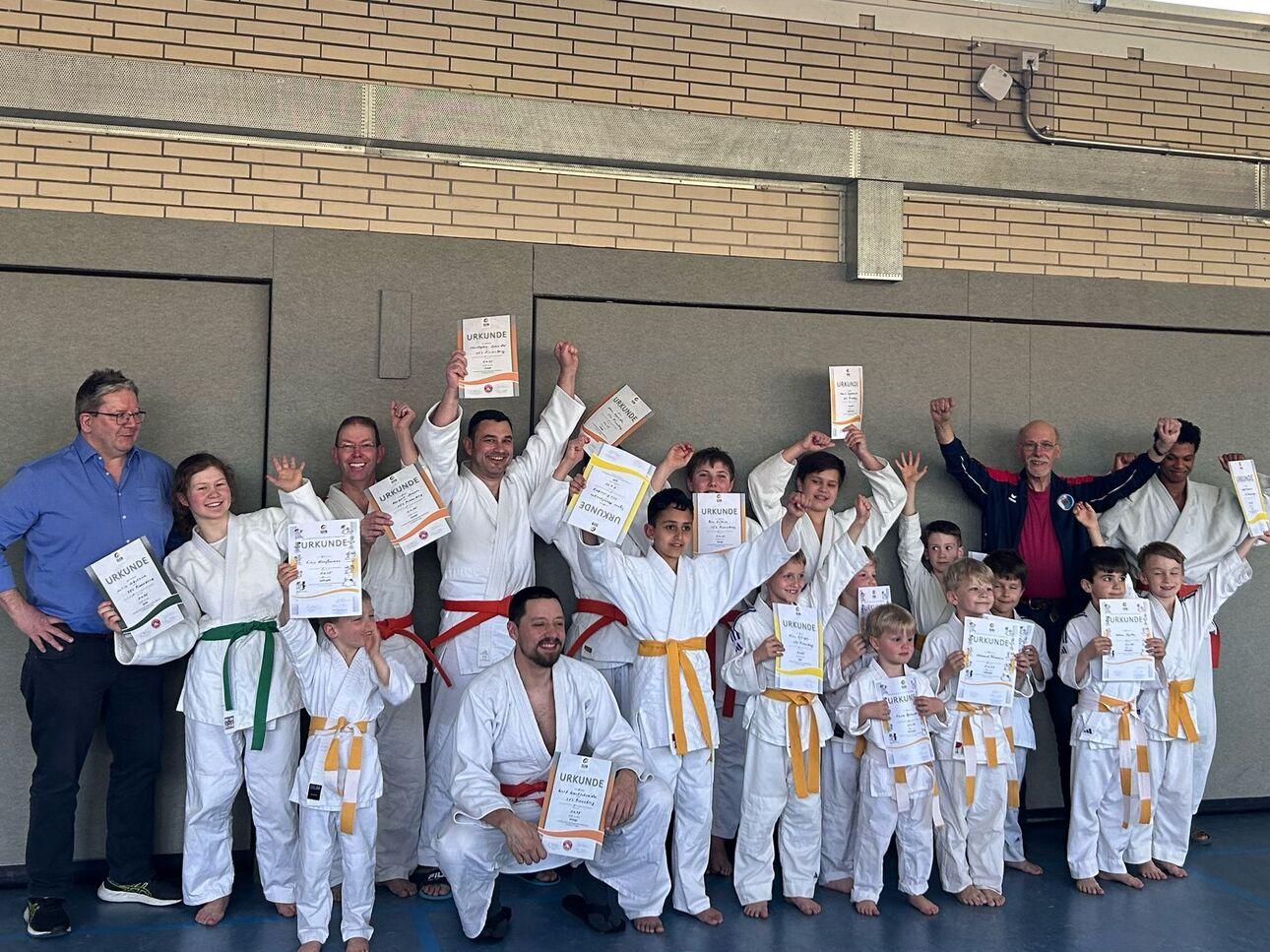 Gruppe von Karatekämpfern in weißen Anzügen mit Urkunden, alle lächelnd und jubelnd in einer Sporthalle.