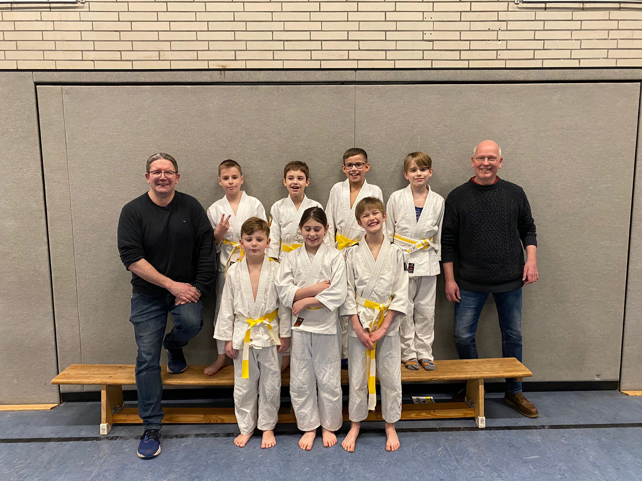 Gruppenfoto von acht Kindern in weißen Karateanzügen mit gelben Gurten und zwei Erwachsenen in einer Sporthalle.