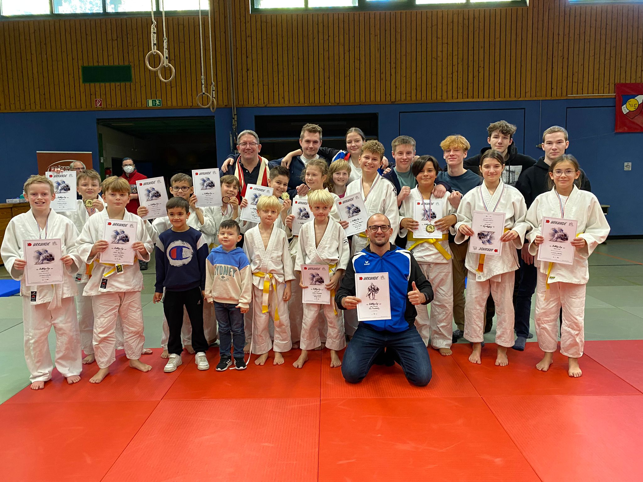 Gruppe von Jugendlichen in Judoanzügen hält Urkunden, umgeben von Trainern in einer Judohalle mit roten Matten.