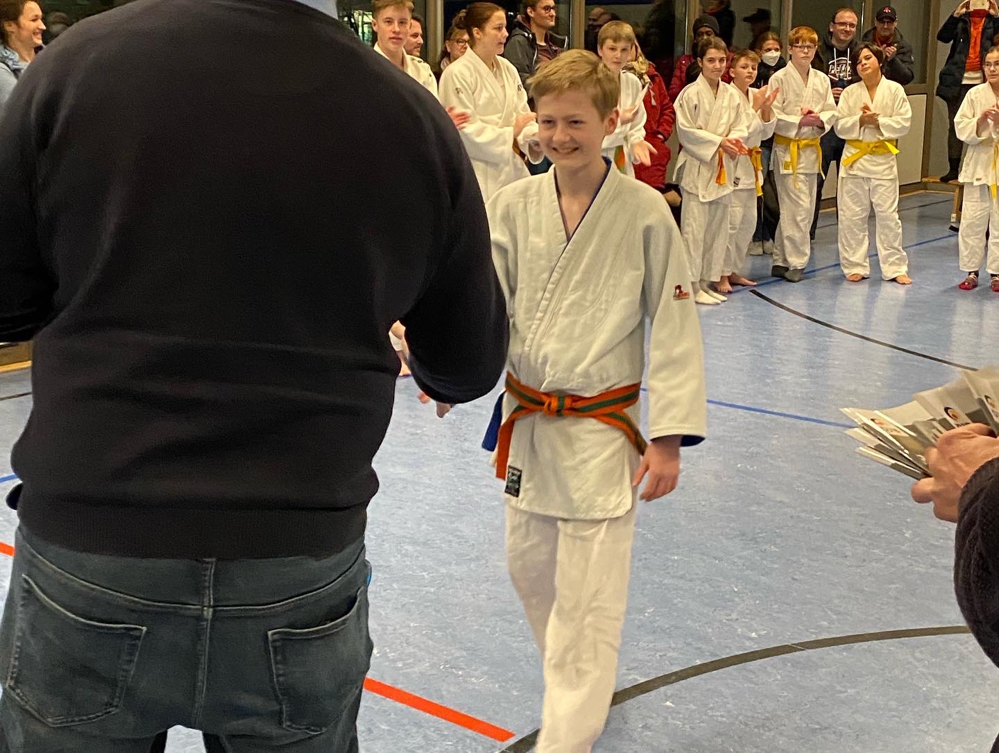 Junge im Karateanzug mit orangem Gürtel schüttelt einem Erwachsenen die Hand, während Zuschauer applaudieren.