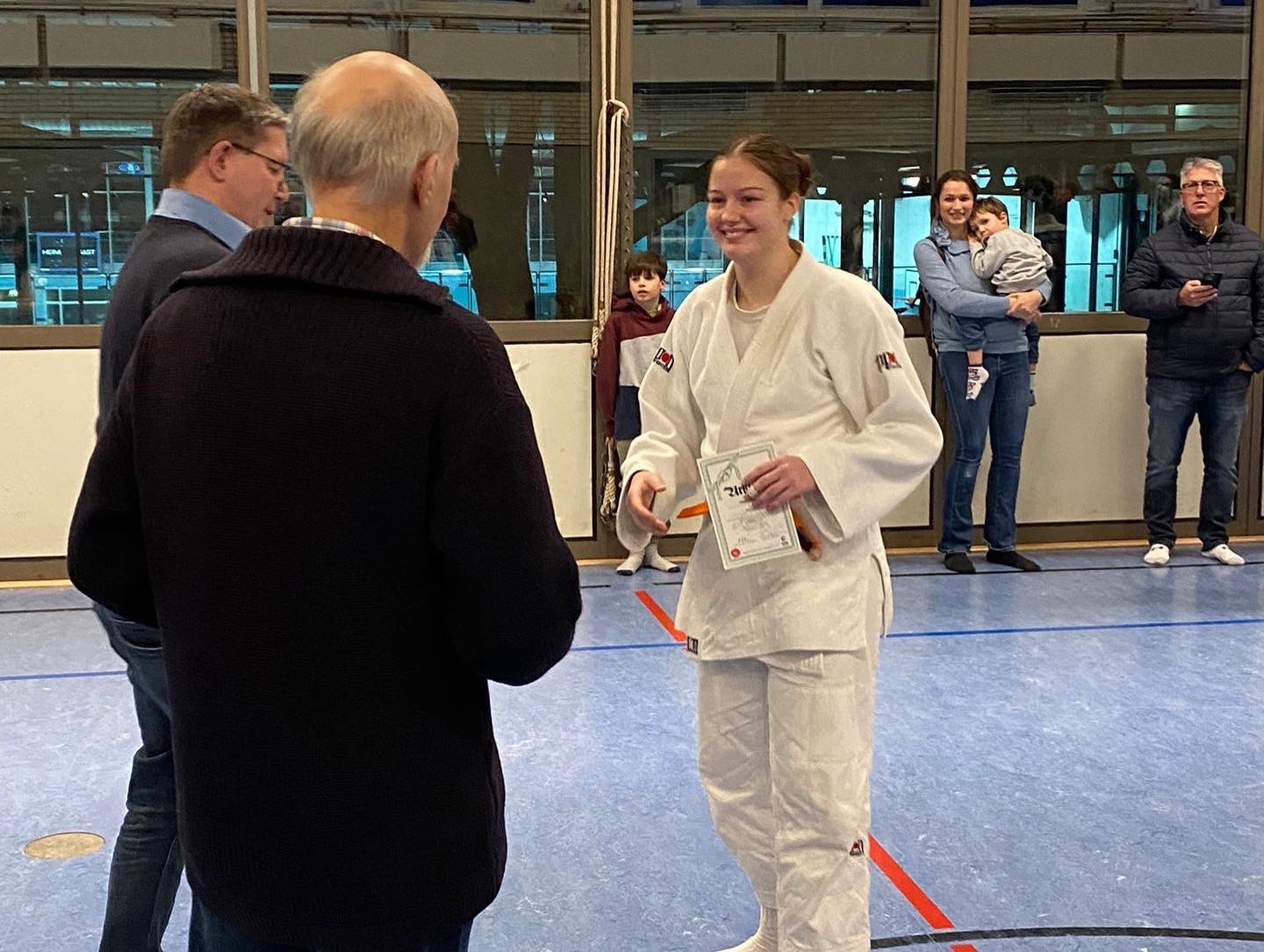 Junge Judoka in weißem Gi erhält stolz ein Zertifikat von einem älteren Mann in einer Sporthalle.