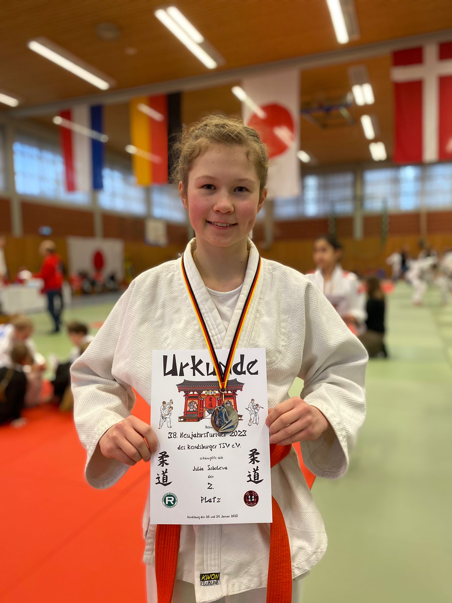 Mädchen in Judogi hält stolz eine Urkunde mit Medaille in einem Sportzentrum, im Hintergrund Flaggen und andere Teilnehmer.