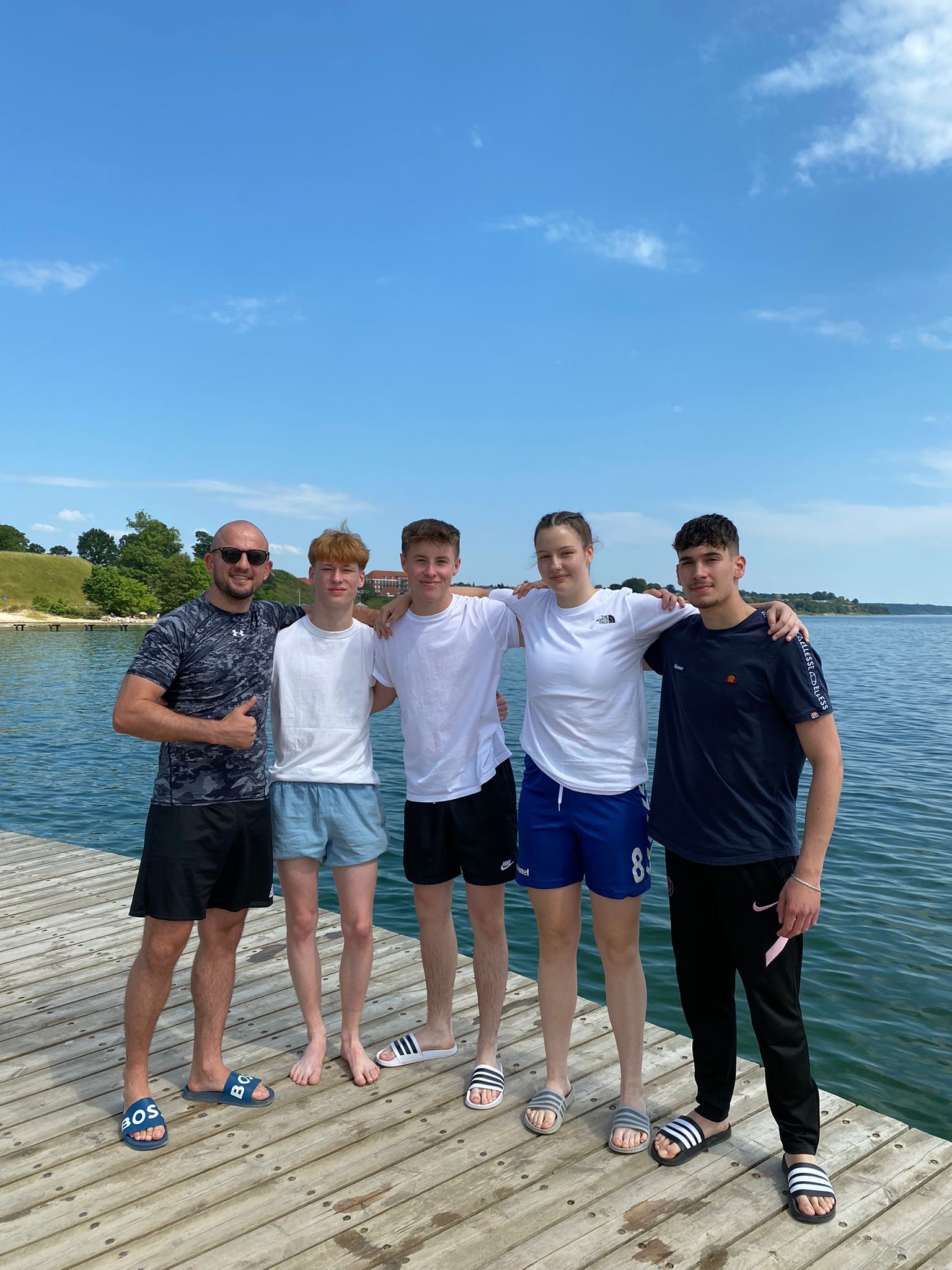 Fünf junge Menschen in sportlicher Kleidung stehen lächelnd auf einem Holzsteg am Wasser unter blauem Himmel.
