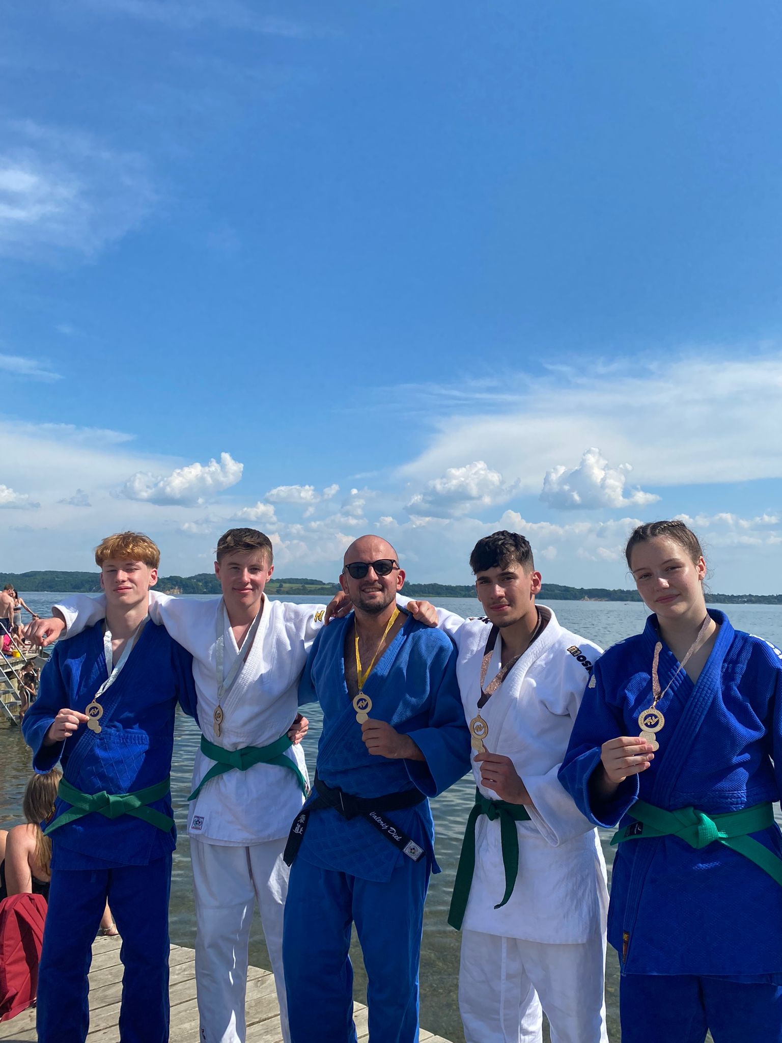 Fünf Judoka in Anzügen mit Medaillen posieren am Ufer eines Gewässers unter blauem Himmel.