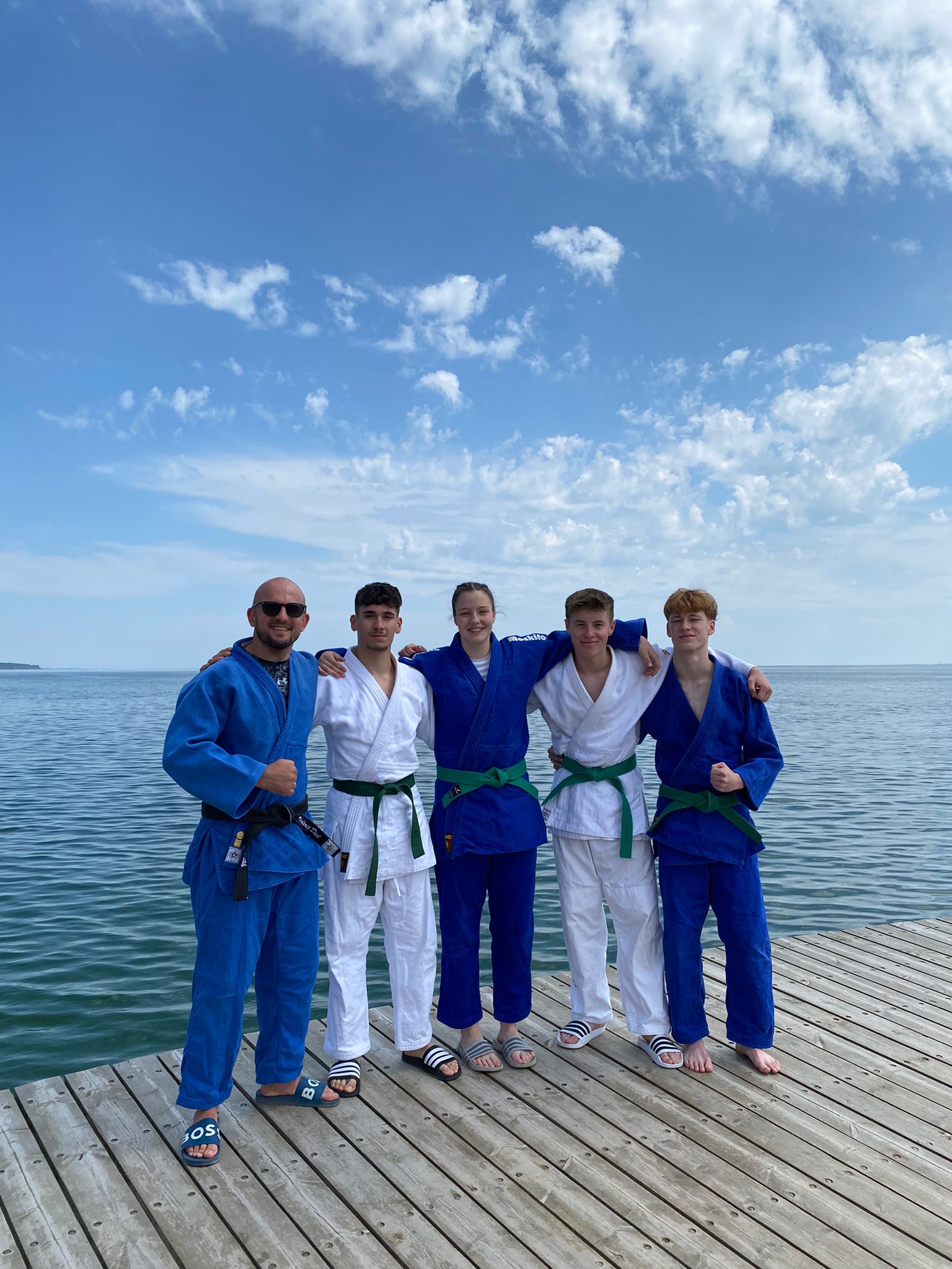 Fünf Personen in BJJ-Anzügen stehen auf einem Holzsteg am Wasser, lächeln in die Kamera unter blauem Himmel.