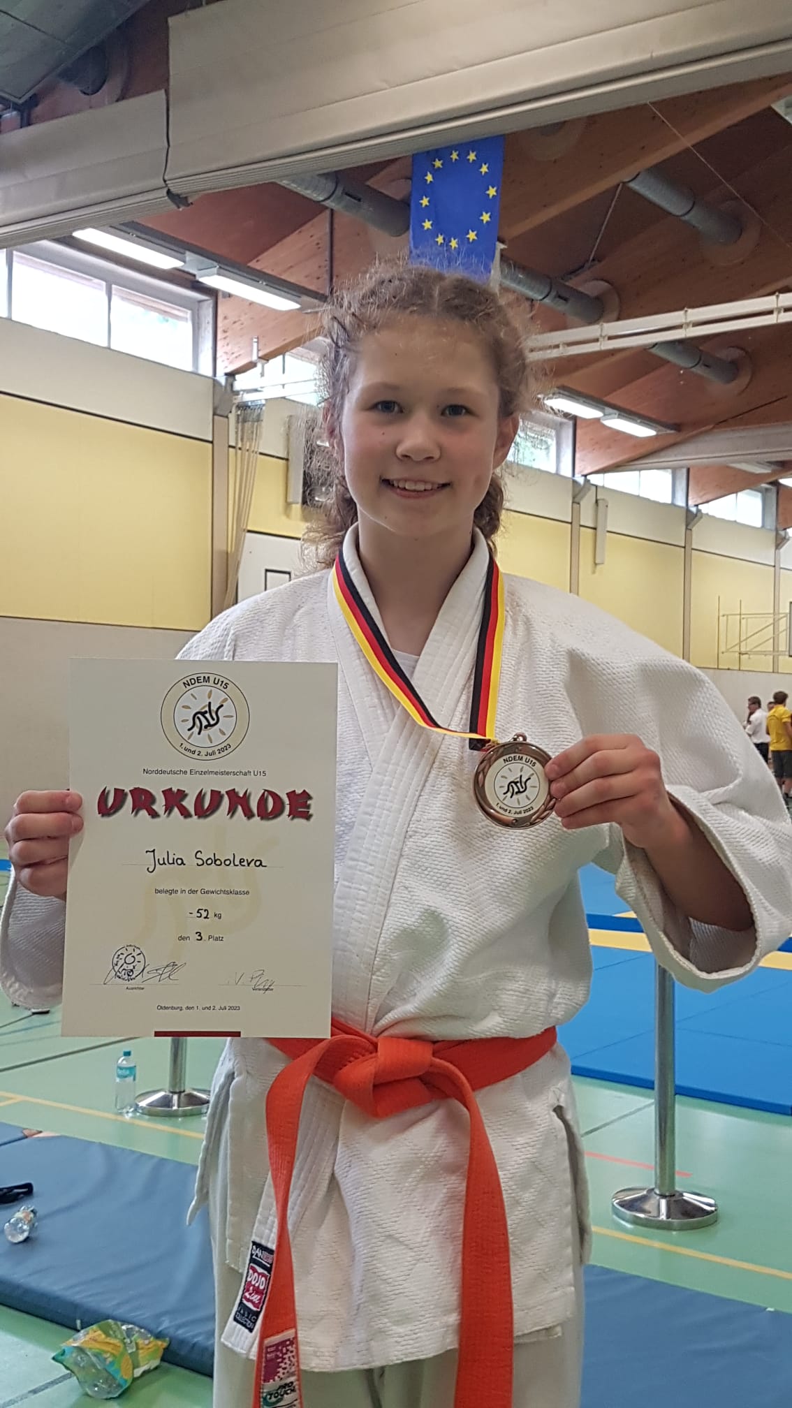 Junge Sportlerin in Judogewand hält Urkunde und Medaille für den 3. Platz in der Gewichtsklasse bis 52 kg.